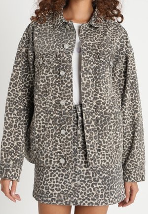 Femme portant une veste en jean gris à imprimé léopard assortie à une jupe avec des boutons argentés, et un haut blanc en dessous.