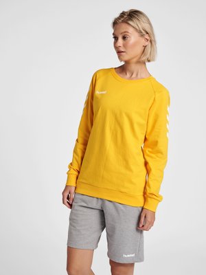 Kvinde iført en gul langærmet Hummel-sweatshirt og grå Hummel-shorts, stående og kigger let til siden mod en hvid baggrund.