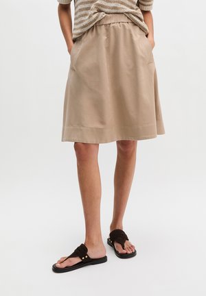 Persoon draagt een beige knielange rok met zakken, een gestreept bovenstuk en zwarte instapsandalen op een effen achtergrond.