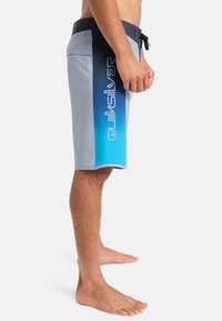 Plavky Quiksilver svetlošedej farby, s modrým gradientným dizajnom a logom. Obsahujú elastický pás a bočnú vrecko.