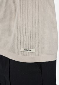 Dobladillo de suéter de punto acanalado beige con etiqueta "PEGADOR" encima del bolsillo de unos pantalones negros.