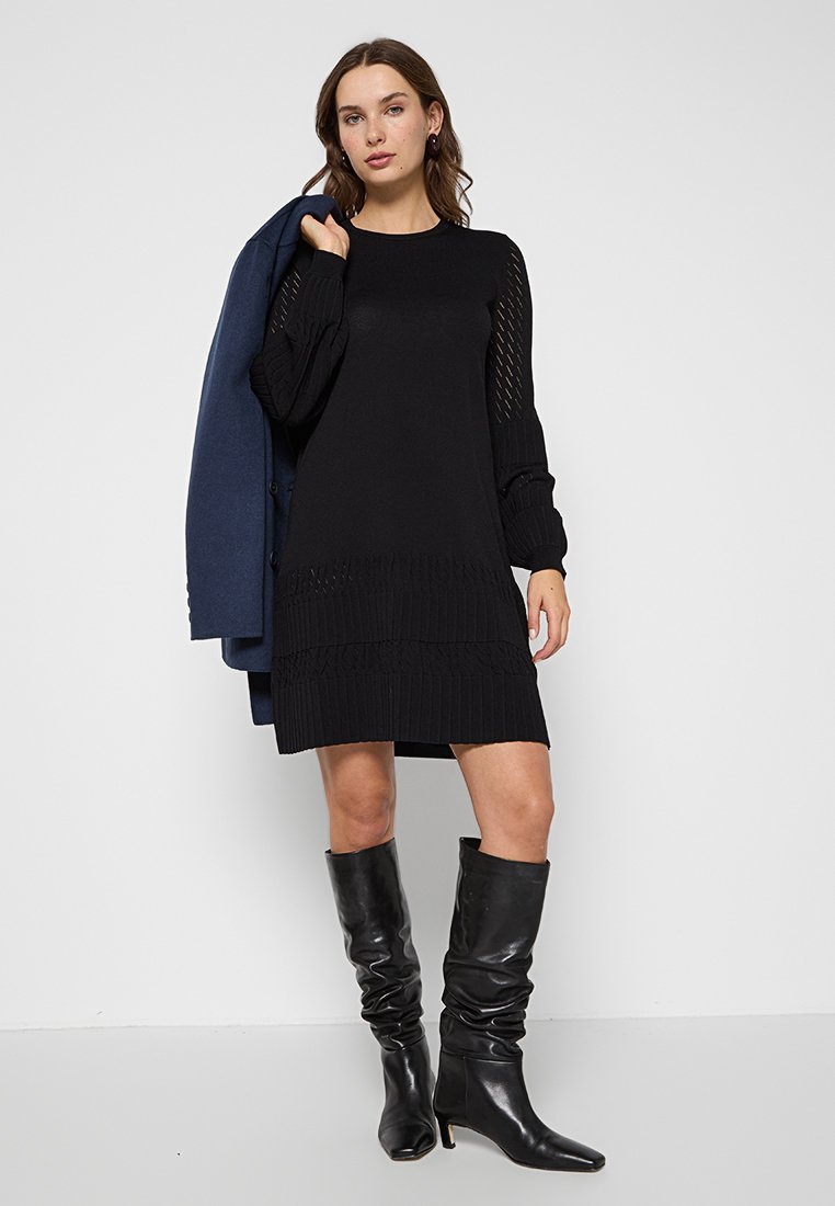 Robe en tricot noir avec texture côtelée, à manches longues et à coupe décontractée, associée à des bottes noires jusqu'aux genoux et à un blazer bleu marine.