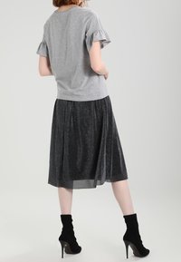 Kvinna står med händerna på höfterna, iförd en grå blus med volanger, svart glittrig midi kjol och svarta högklackade ankelskor.