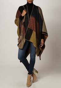Multi-colored poncho featuring geometric patterns in brown, navy, and olive, with fringed edges. Paired with fitted jeans and beige ankle boots.