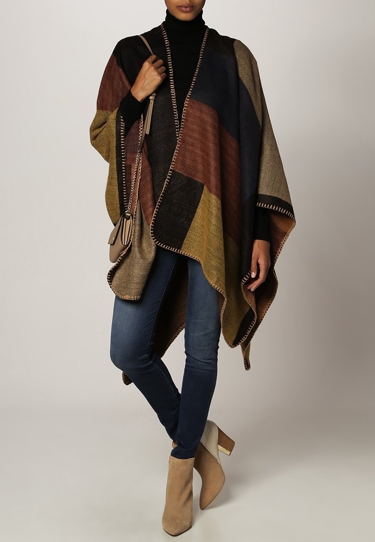 Multi-colored poncho featuring geometric patterns in brown, navy, and olive, with fringed edges. Paired with fitted jeans and beige ankle boots.