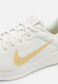 Hvide stof sneakers med en tekstureret overdel, guld Nike swoosh logo, flad hvid sål og beige tungepatch. Snørebånd i matchende hvid.