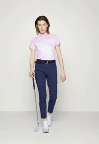 Polo majica s kratkimi rokavi v roza tie-dye vzorcu, mornarsko modri golf hlače slim-fit, črn pas, drži golf palico, bele športne čevlje.