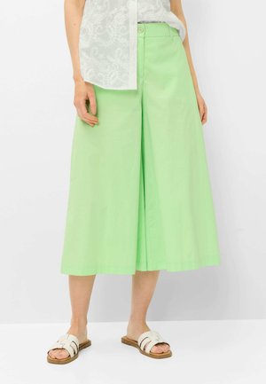 Vrouw draagt lichtgroene wijde cropped broek, witte kanten top en witte platte slides, staand tegen een effen achtergrond.