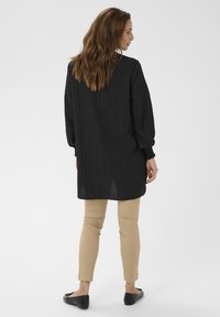 Dlhá tunika s čiernymi pruhmi a rozšírenými rukávmi, v kombinácii s béžovými úzkymi nohavicami a čiernymi plochými topánkami, z pohľadu zozadu.