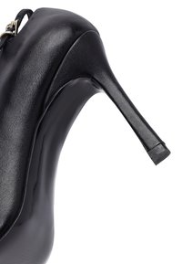 Zapato negro de cuero de tacón alto con un delgado tacón de aguja y una correa en el tobillo con una hebilla de metal, mostrado desde un lado.