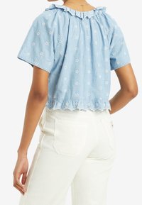 Haut bleu chambray à manches courtes avec broderies florales blanches, décolleté froncé et ourlet festonné. Associé à un pantalon blanc taille haute.