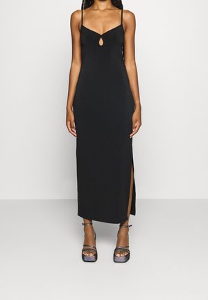 Femme portant une robe longue noire sans manches avec de fines bretelles, un détail œillet devant, une fente latérale et des sandales à plateforme métalliques.