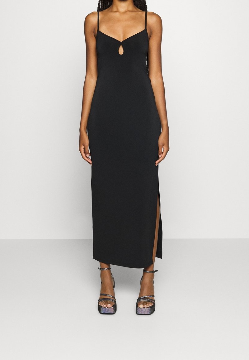 Femme portant une robe longue noire sans manches avec de fines bretelles, un détail œillet devant, une fente latérale et des sandales à plateforme métalliques.
