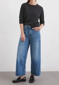 Osoba v černém dlouhém rukávu, volných modrých džínách s vysokým pasem, černých mokasínech, s jednou rukou v kapse, stojící na obyčejné podlaze.