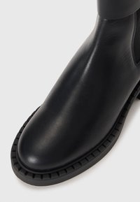 Bottine en cuir noir, à bout arrondi, avec une surface texturée et une semelle en caoutchouc présentant un détail à bord ondulé.