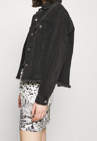 Veste en denim noire avec fermeture à boutons, poches poitrine et ourlet effiloché. Associée à une jupe blanche à motifs avec des motifs de plantes.
