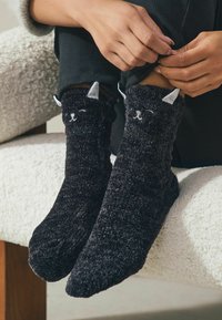 Svarta, fluffiga strumpor med en fantasifull kattansiktsdesign, som har vita öron och broderade detaljer. Mjuk textur, ankellängd och en bekväm passform.