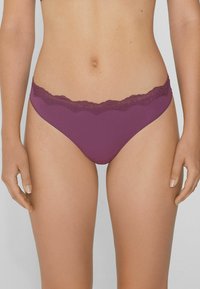 Kobieta w purpurowej bieliźnie w stylu bikini z koronkowym wykończeniem, stojąca z ramionami swobodnie opartymi wzdłuż ciała na neutralnym tle.