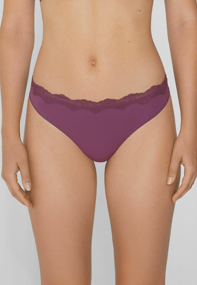 Kobieta w purpurowej bieliźnie w stylu bikini z koronkowym wykończeniem, stojąca z ramionami swobodnie opartymi wzdłuż ciała na neutralnym tle.