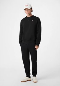 Sort sweatshirt og matchende bukser lavet af stof, med en lille logo på brystet. Suppleret med en hvid kasket og cremefarvede sneakers.