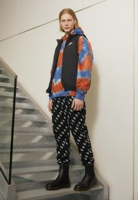 Crni prsluk od pufinga preko dukserice s multikolor tye-dye uzorkom u plavoj i narančastoj boji. Crne trenirke s bijelim tekstom "JORDAN". Crne gležnjače. Sive stepenice.