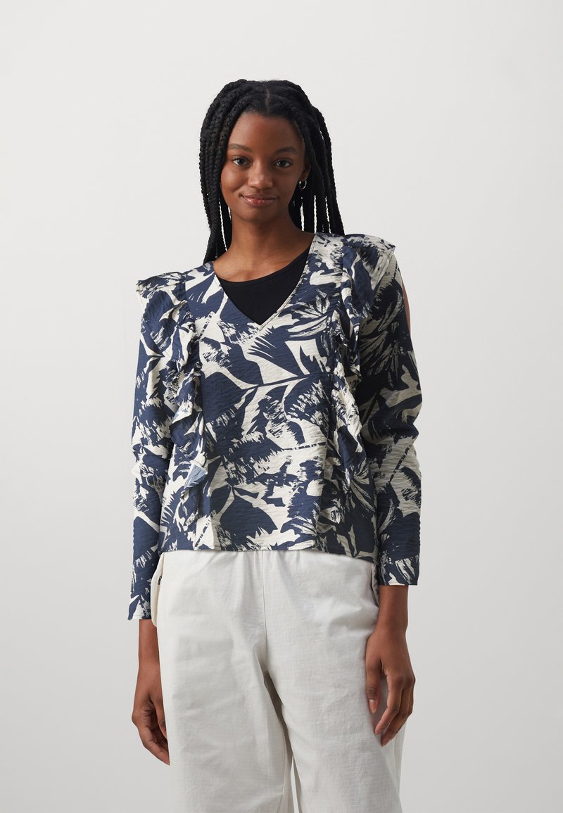 Maglietta a maniche lunghe con un motivo astratto blu navy e crema, con spalle a volant e scollo a V, abbinata a pantaloni bianchi.