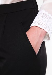 Svarta skräddarsydda byxor med sidoficka och en texturerad vit spetsärm från en skjorta synlig vid manschetten. Slät tygfinish.