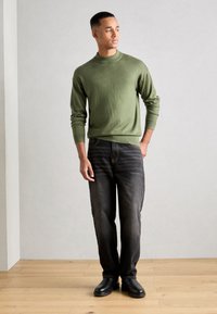 Pulover verde din tricot, cu guler rotund, asortat cu blugi negri decolorati și pantofi negri. Puloverul are mâneci lungi și manșete cu nervuri.
