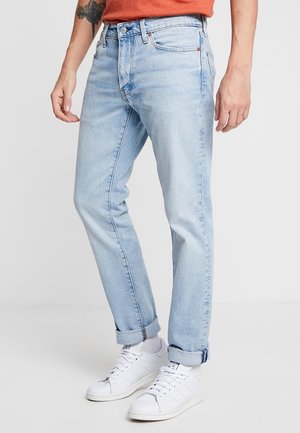 Homme portant un jean bleu clair retroussé, des chaussettes blanches, des baskets blanches et une chemise orange avec un tatouage sur son avant-bras gauche.