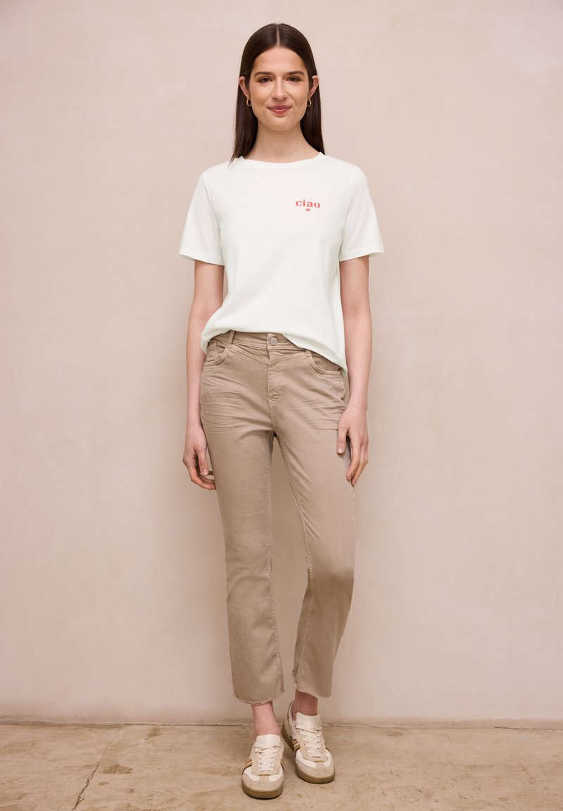 Jeune femme portant un t-shirt blanc avec le texte "ciao", pantalon beige et baskets blanches, debout devant un mur beige uni.