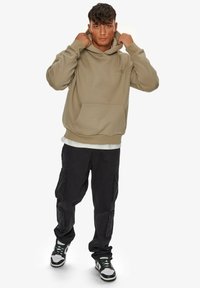 Tan hoodie med en frontlomme og justerbar snor, parret med sorte cargo-bukser. Modellen har sorte og hvide sneakers på.