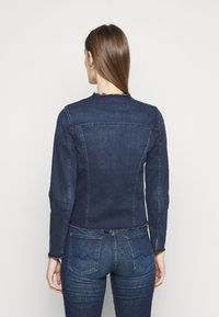 Chaqueta de mezclilla azul oscura con un diseño ajustado, dobladillo sin acabar y mangas largas. Presenta costuras visibles y un yugo trasero.