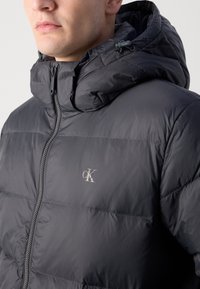 Gevoerde donkergrijze jas met een ritssluiting en een hoge kraag. Bevat een opvallend zilverlogo op de borst en een verstelbare capuchon.