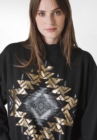 Svart sweatshirt med ett geometriskt mönster i guld och silver. Den har en hög krage och en mjuk textur, med mönster som inkluderar pilar och diamanter.