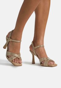 Beżowe sandały na wysokim obcasie z błyszczącymi akcentami z cyrkonii, krzyżującymi się paskami i prostokątnym obcasem. Miękka zamszowa struktura.