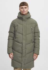 Polstret parka i olivengrønn med høy krage, glidelåslommer foran og lang design. Glatt tekstur og horisontale quiltedetaljer.