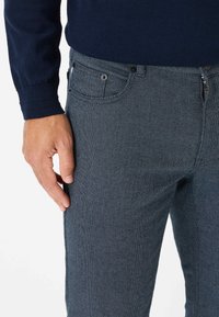 Donkerharingbone mannenbroek met een ritssluiting, zijzakken en een subtiel gestructureerd patroon. Hand rustend op de broekstof.