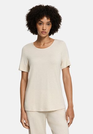 Vrouw met krullend haar draagt een beige top met korte mouwen en bijpassende broek, staand tegen een effen witte achtergrond.