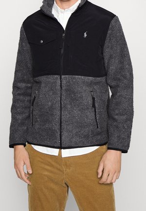 Mies, joka pitää mustaa ja harmaata vetoketjullista fleecetakkiin, jossa on rintatasku, ja vaaleanruskeita samettihousuja, seisoo yksinkertaisen taustan edessä.