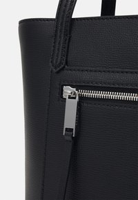 Musta teksturoidulla nahkatote laukkumalli, jossa on hopeinen vetoketju ja pieni nahkavetoketjun vedin. Täydentää ommellut yksityiskohdat ja tukevat kahvat.