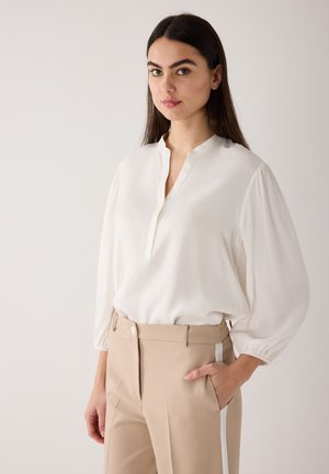 Woman with long dark hair wearing a white blouse with puffed sleeves and beige trousers with a white side stripe, standing with one hand in pocket.
