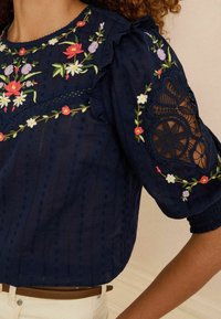 Navy blue blouse with floral embroidery, textured fabric, ruffled neckline, and lace details on sleeves. Fitted with elastic cuffs.