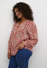 Blusa de patrón de hojas en rojo y blanco con escote en V, mangas largas y un ajuste relajado, combinada con jeans de mezclilla azul.