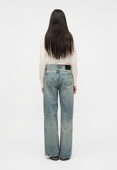 Άτομο με μακριά σκούρα μαλλιά, φορώντας μπεζ πουλόβερ και ξεθωριασμένο μπλε τζιν με φαρδιά πόδια, στέκεται με την πλάτη γυρισμένη στην κάμερα σε απλό φόντο.