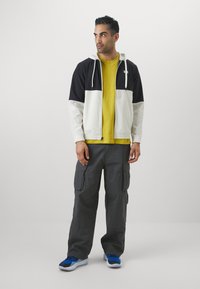 Giacca con cappuccio con pannelli neri e crema, t-shirt gialla, pantaloni cargo grigi con tasche e scarpe blu, in piedi su uno sfondo neutro.