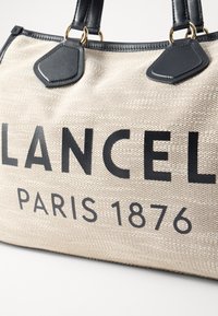 Beige plátěná taška s černými koženými akcenty, s velkým černým nápisem "LANCEL PARIS 1876". Obsahuje zlaté kovové detaily a dvě ucha.