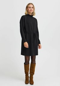 Crna haljina dugih rukava s lažnim turtleneck ovratnikom, pojasom na vezanje i opuštenim krojem. Nosena s bež čizmama do koljena i crnim tajicama.