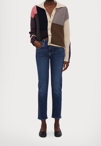 Multikleurige cardigan met een kraag, met verschillende breipatronen, knopen en blauwe skinny jeans gecombineerd met zwarte flats.