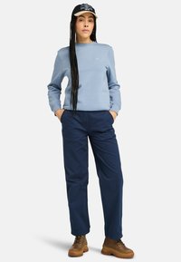 Felpa azzurra con scollo rotondo, pantaloni blu navy e stivali marroni con lacci. Il modello ha lunghe trecce e un cappellino nero con dettaglio del logo.