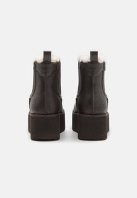 Botines de color marrón oscuro con tacón cuadrado, patrón geométrico en relieve y forro de piel sintética en el cuello. Logo de GUESS en la parte posterior.